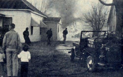A historical photo of a house burning in the background with an old fire truck in the foreground and firefighters spraying water on the fire